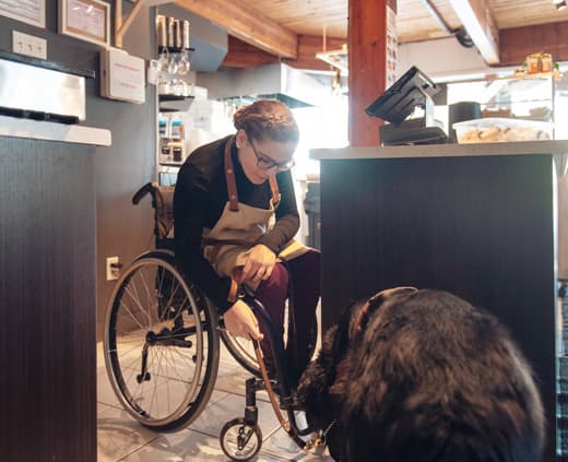 Une employée d’une boutique dans un fauteuil roulant se penche pour interagir avec son chien d’assistance. Elle porte des lunettes et un tablier.